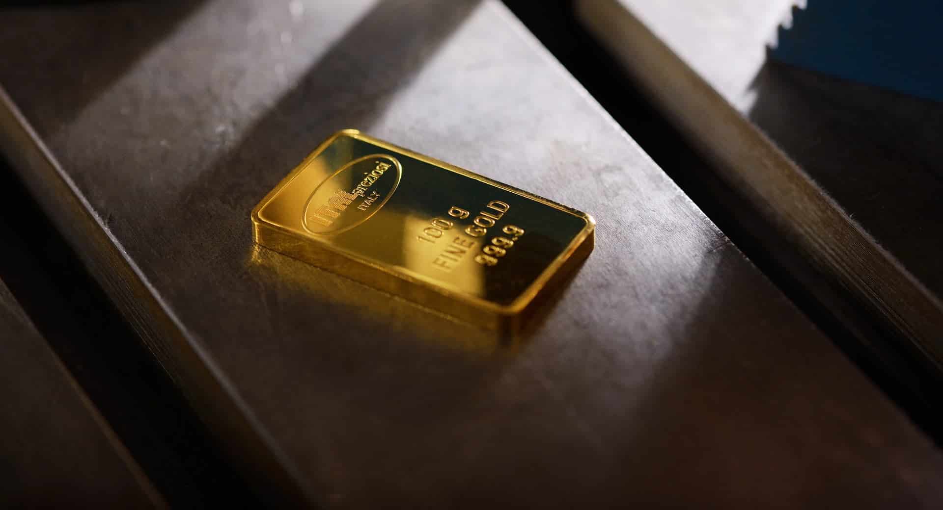 Un singolo lingotto d'oro del peso di 100 grammi poggia su una superficie metallica scura, riflettendo la luce e mostrando incisioni che ne indicano il peso e la purezza.