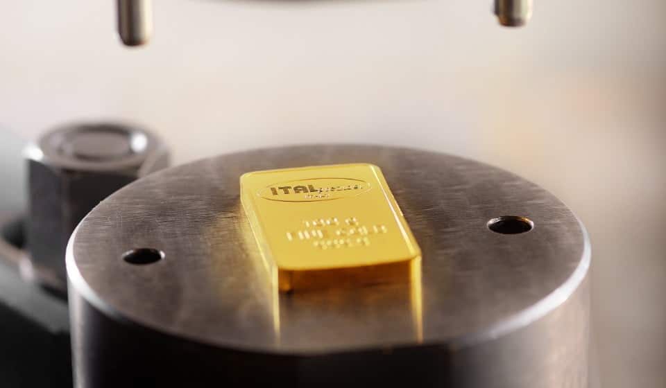 Un lingotto d'oro timbrato con la scritta ITALPREZIOSI poggia su una piattaforma metallica rotonda, con parte di un macchinario visibile sopra e a lato, che suggerisce un processo di saggiatura o di analisi.