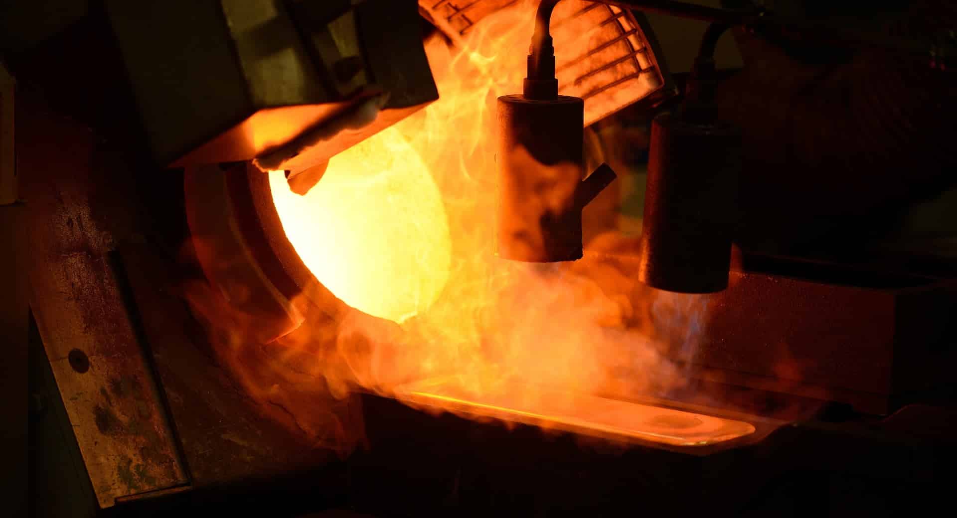 Il metallo fuso viene versato da una fornace in uno stampo, con una luce arancione brillante e il vapore o il fumo in aumento che creano una drammatica scena industriale.